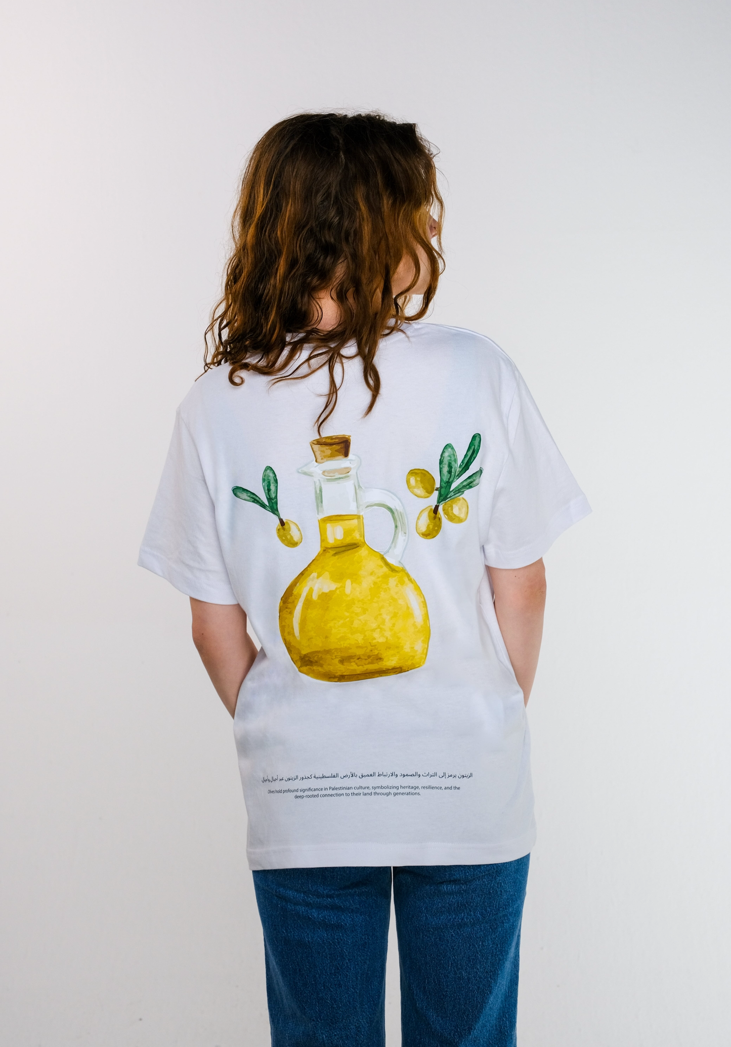 Back view of the model wearing the Land of Olives T-shirt, showing a hand-painted-style illustration of an olive oil bottle surrounded by olives, with Arabic and English text highlighting the deep connection between Palestinian heritage and olive trees.