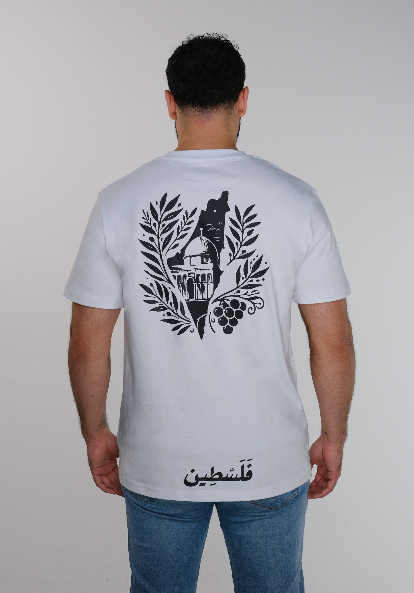 A model wearing the T-shirt from the back, showcasing a stylized map of Palestine framing Al-Aqsa Mosque, surrounded by olive leaves, with a grape cluster on the right side and "فلسطين" written in Arabic at the bottom which mean "Palestine".