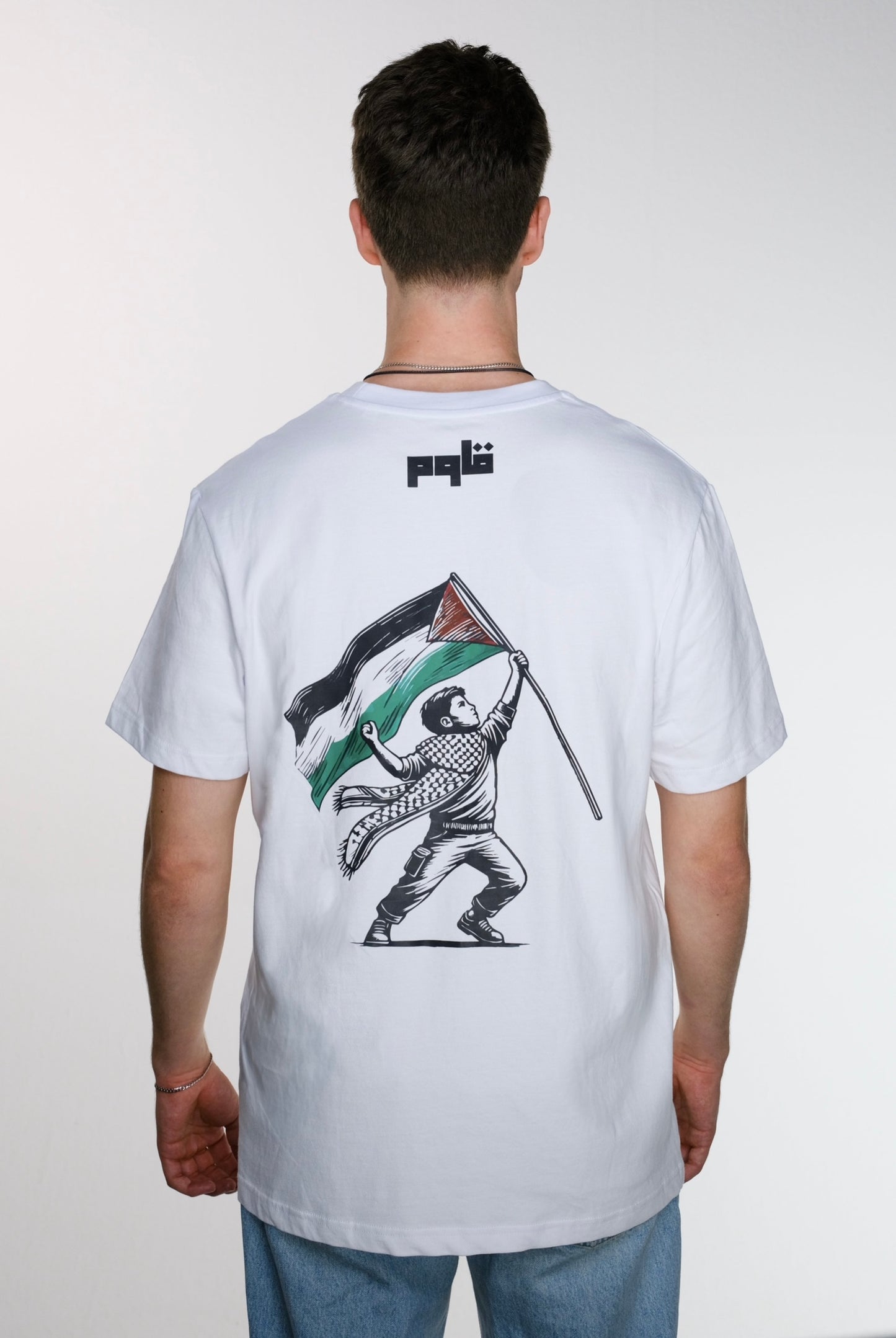 Model wearing a Resist T-shirt, Back view of a white Resist T-shirt, showing Arabic word "قاوم" at the top which mean Resist, and a graphic of a child holding a Palestinian flag in one hand and appearing to throw something with the other, wearing a traditional keffiyeh—symbolizing resistance and Palestinian identity.