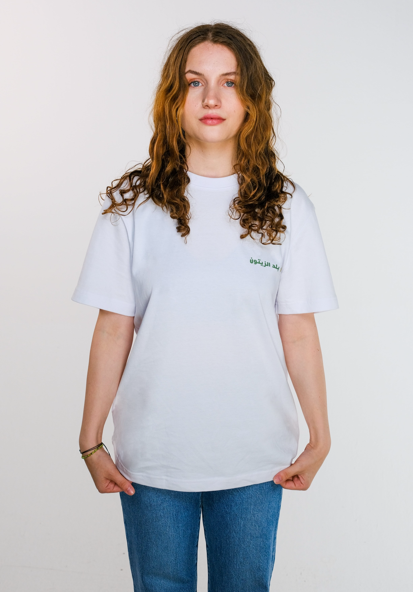 The model holding the edges of the Olive Land T-shirt to show the soft cotton fabric, highlighting the front chest design of olive leaves, twig and Arabic text.