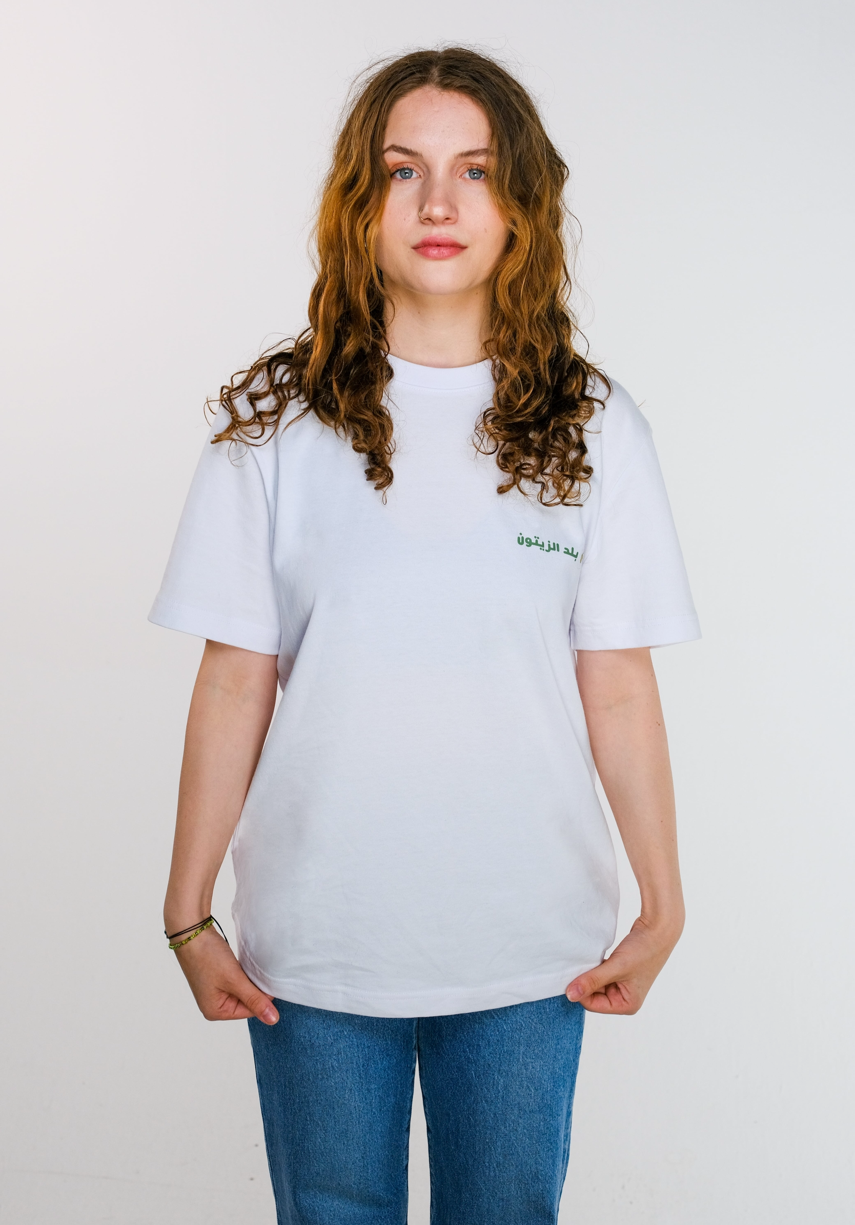 The model holding the edges of the Olive Land T-shirt to show the soft cotton fabric, highlighting the front chest design of olive leaves, twig and Arabic text.