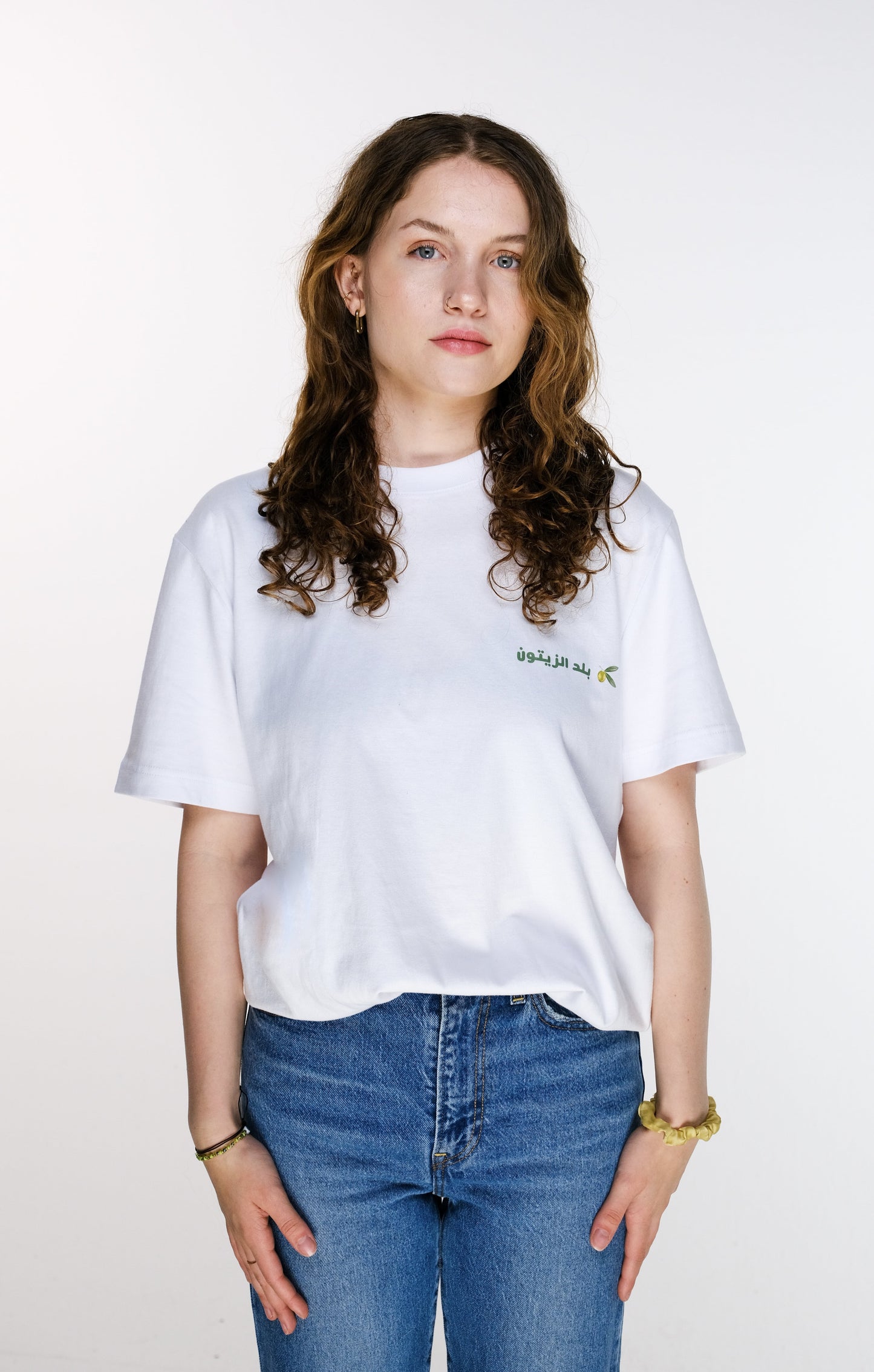 Front view of the model wearing the Land of Olives T-shirt, with the shirt neatly tucked into the pants. The design features a small green Arabic calligraphy reading “بلد الزيتون” (Land of Olives) alongside a delicate olive twig near the chest.