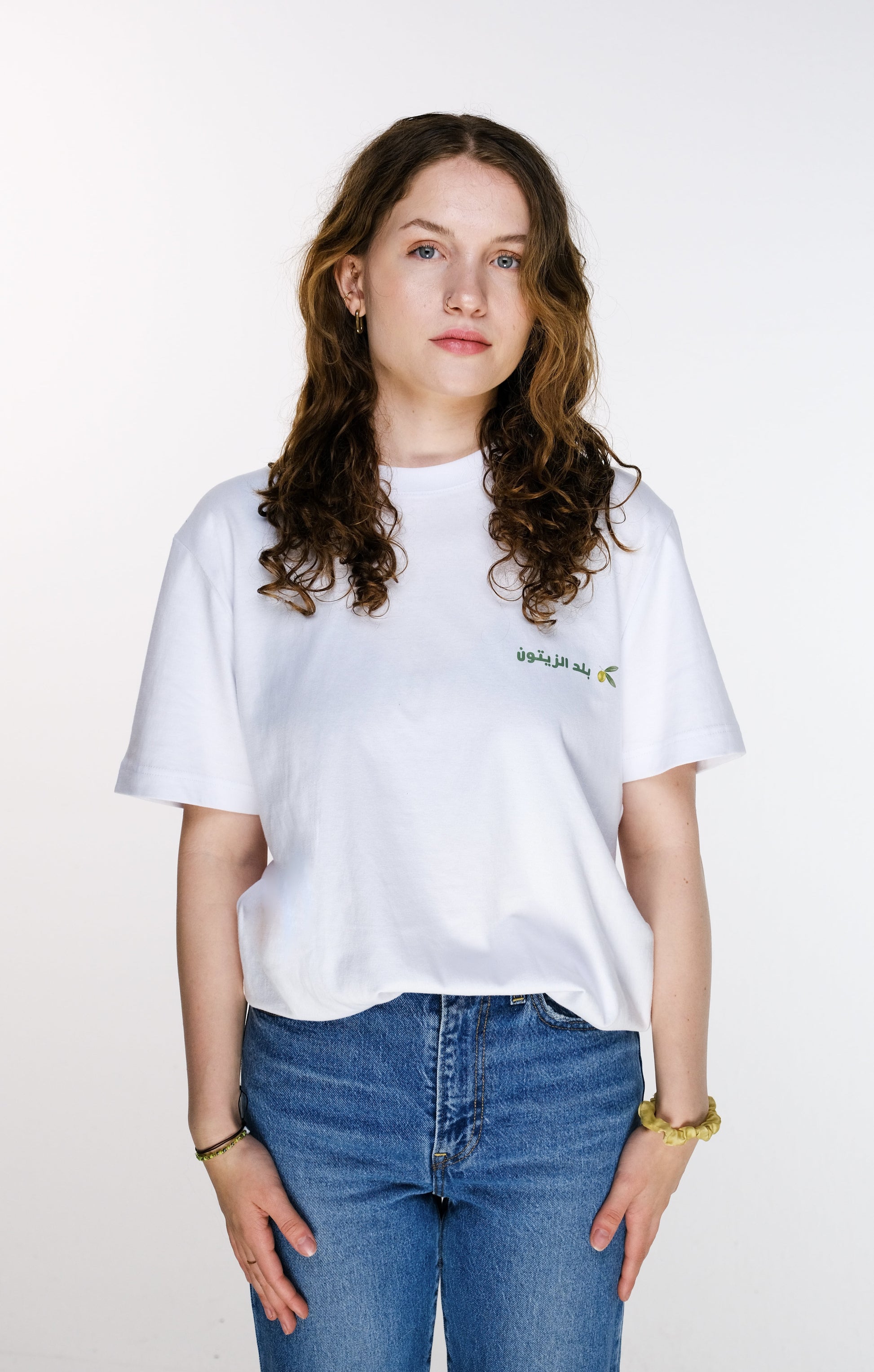 Front view of the model wearing the Land of Olives T-shirt, with the shirt neatly tucked into the pants. The design features a small green Arabic calligraphy reading “بلد الزيتون” (Land of Olives) alongside a delicate olive twig near the chest.